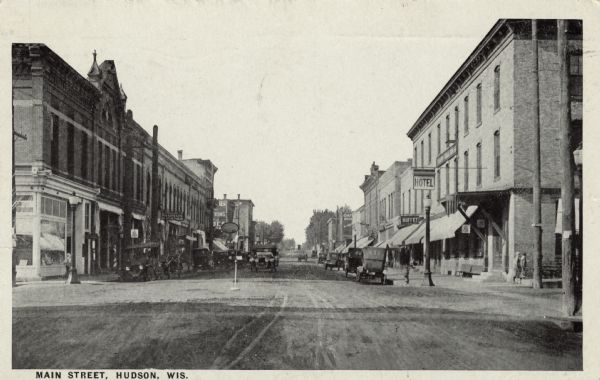 View down unpaved Main Street, a central business district. Businesses include a pharmacy, a buffet, and a hotel. Automobiles are parked along the curbs. Caption reads: "Main Street, Hudson, Wis."