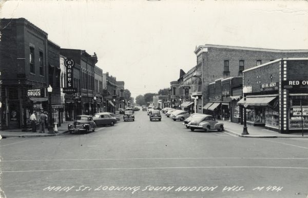 View looking south down Main Street, which is lined with commercial business. Automobiles are parked at an angle on both sides of the street. Caption reads: "Main St. Looking South, Hudson, Wis."