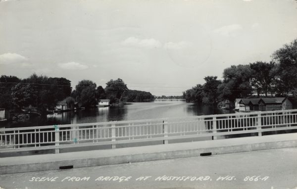 View of the Rock River from a bridge at Hustisford. The river is lined with boathouses. Caption reads: "Scene from Bridge at Hustisford, Wis."
