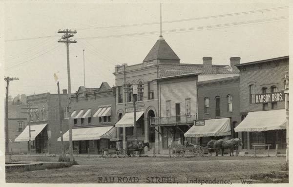 View across unpaved intersection towards businesses, including the A. Garthus building with a grocery/general store, the State Bank of Independence, the Thos. E. Lokken bar or restaurant, the Independence Drugstore and Red Cross Pharmacy, and Hanson Bros. Horse-drawn vehicles are at the curb.