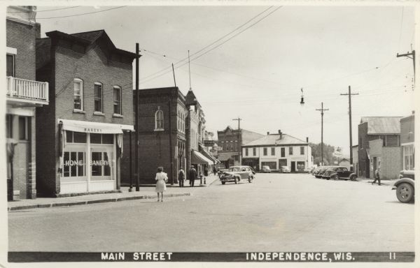 View down Main Street, with the Home Bakery on the left at the street corner. Further up the block on the left are pedestrians on the sidewalk, and signs on storefronts that read: "Drugs Soda," "Philco Radio" and "Cafe." Automobiles are parked at an angle along the curbs.