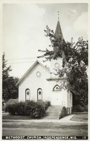 View across street towards a white wooden church with stained glass windows. There are stairs on the right leading up to the entrance, and a steeple above it. Caption reads: "Methodist Church, Independence, Wis."