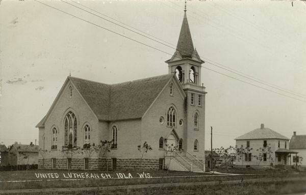 Three-quarter view of front and left side of the Lutheran Church. There are stairs leading up to the entrance, and a bell tower is on the right. The brick church has gothic arches and stained glass windows. Houses are in the background. Caption reads: "United Lutheran Ch. Iola, Wis."