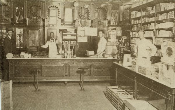 Interior view of a meat market, with four men standing behind the counters. Deer head trophies are mounted on the back wall. Caption on back reads, in part: "Note the elaborate woodwork on the cooler."