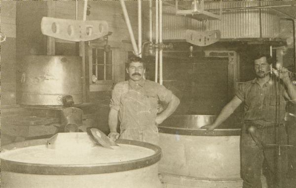 Interior view of a cheese factory, with two men posing. Caption on back reads in part: "Cooking the milk in copper-lined vats."