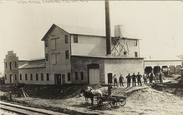 Slightly elevated view of a canning factory, with workers lined up out front, and a man standing in a wagon pulled by a team of horses. Railroad tracks are in the foreground. Caption reads: "Iron Ridge Canning Factory, Wis."