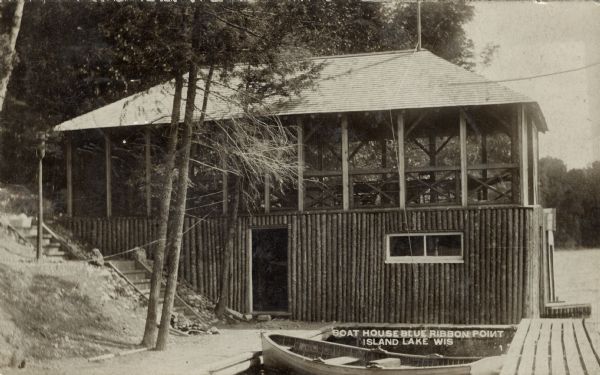 Photographic postcard view of a wooden boathouse at the shoreline, on the slope of a wooded hill. There is a porch on the upper level. A rowboat is moored at the dock in the foreground. Caption reads: "Boat House, Blue Ribbon Point, Island Lake, Wis."