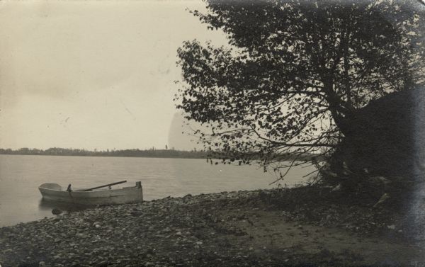 View of a lake shore with a rowboat pulled up to a stone beach. There are trees on the right, and the far shoreline is in the distance.