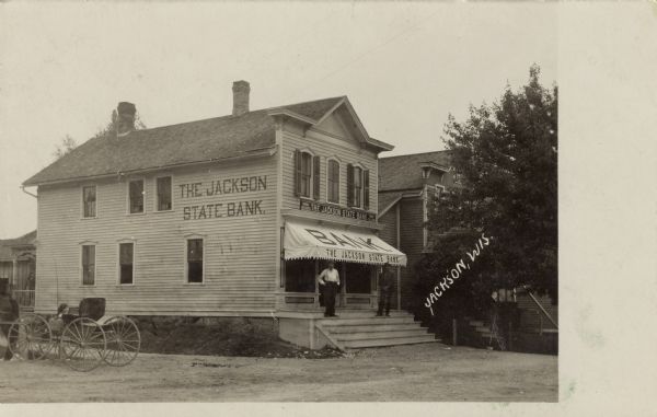 Black and white photographic view of the bank building. Two men are standing on the front steps, and a buggy parked on the left side. Caption reads: "Jackson, Wis."