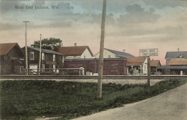 View of Jackson from across the railroad tracks. The depot is on the left. A "Look Out for Cars" sign is at the railroad crossing. Caption reads: "West End Jackson, Wis."