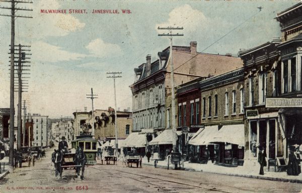 View of Milwaukee Street in central Janesville lined with businesses. Horse-drawn vehicles and a streetcar are in the street. Caption reads: "Milwaukee Street, Janesville, Wis."