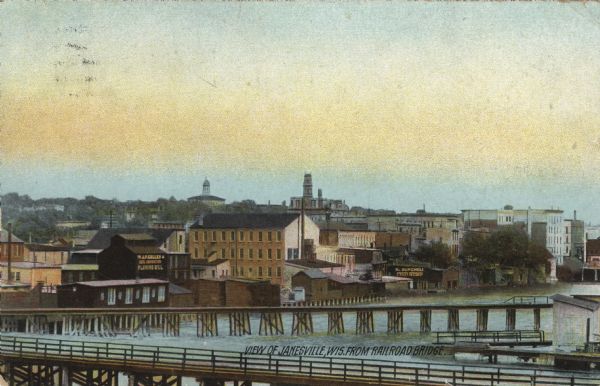 Colorized postcard view of central Janesville from the railroad bridge over the Rock River. Warehouses are along the river's edge. Caption reads: "View of Janesville, Wis. from Railroad Bridge."