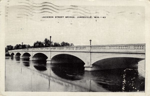 View across water toward a concrete arch bridge over the Rock River. Caption reads: "Jackson Street Bridge, Janesville, Wis."