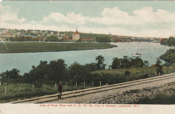 Elevated view of the Rock River. In the foreground is a man standing on railroad tracks. Janesville is on the far shoreline. There is a bridge in the distance on the right, and an excursion boat is on the river. Caption reads: "View of Rock River and C. N. W. Ry. City in distance, Janesville, Wis."