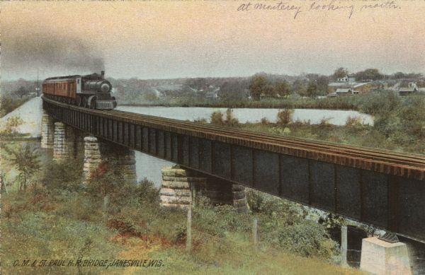 Elevated view of a curving railroad bridge with a train approaching. The Rock River is below. Buildings are on the far bank on the right. Caption reads: "C. M. & St. Paul R. R. Bridge, Janesville, Wis."