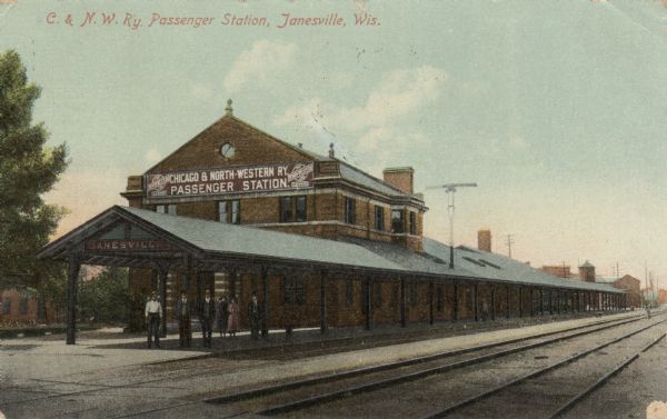 View across railroad tracks toward the station, with a few passengers on the platform. Caption reads: "C. & N. W. Passenger Station, Janesville, Wis."