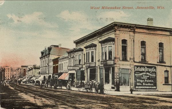 View of buildings along one side of a downtown street. Streetcar tracks run along the street. There is a large sign on the side of a building on the right for "Reliable Drug Co." Caption reads: "West Milwaukee Street, Janesville, Wis."