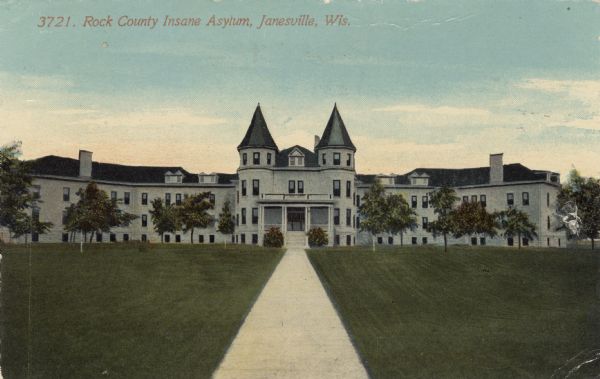 View up sidewalk toward the insane asylum. Turrets are on either side of the entrance, and wings with wards are on both sides. Caption reads: "Rock County Insane Asylum, Janesville, Wis."