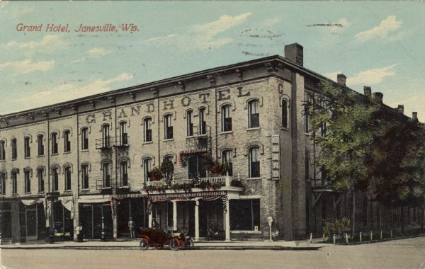 View from intersection towards a hotel on the corner, with an electric sign. An automobile is parked at the curb, and a public scale is on the sidewalk. Caption reads: "Grand Hotel, Janesville, Wis."