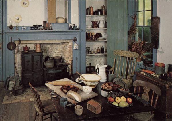 View of Tallman House kitchen. There is a wood-burning stove in the fireplace. Fruit and nuts are arranged on the table.