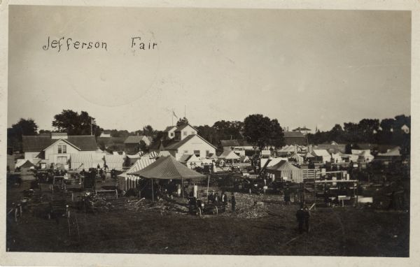 Elevated view of a community fair with tents set up for exhibits. Two buildings with signs are just beyond the tents on the midway, with signs that read, on the left: "Art", and another building on the right that reads: "Industrial". Caption reads: "Jefferson Fair."