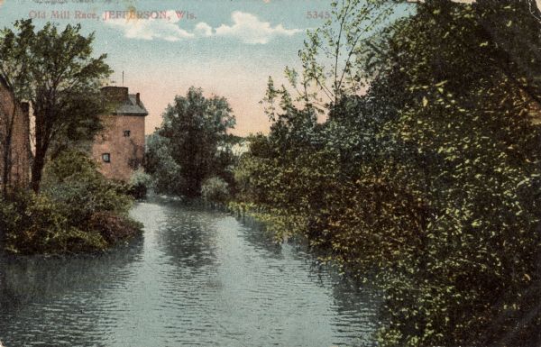 View down a tree-lined river toward a mill in the background. Caption reads: "Old Mill Race, Jefferson, Wis."