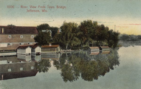View across a still river with buildings reflected in it. Boathouses are along the shore. Caption reads: "River View from Town Bridge, Jefferson, Wis."