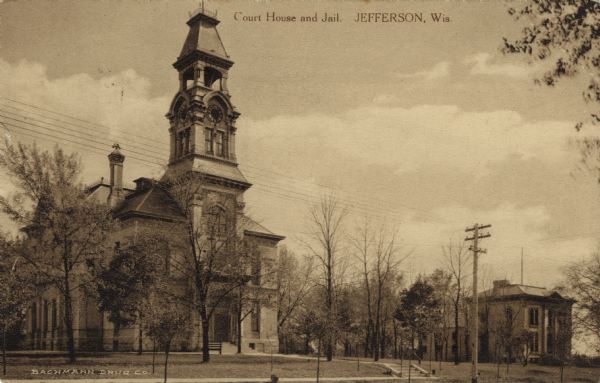 View of the courthouse with a clock tower above the entrance. Saplings are on the lawn. Caption reads: "Court House and Jail, Jefferson, Wis."