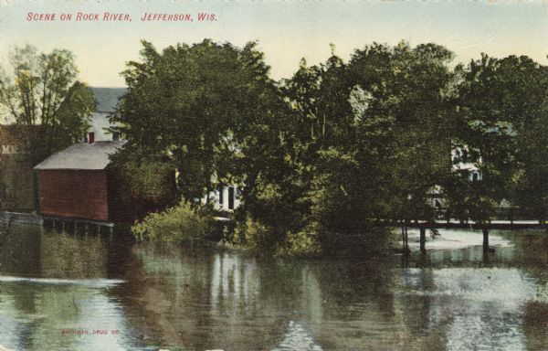 View across water toward a bend in the Rock River with a footbridge crossing it on the opposite shoreline. Buildings are along the shoreline and behind the trees. Caption reads: "Scene on Rock River, Jefferson, Wis."