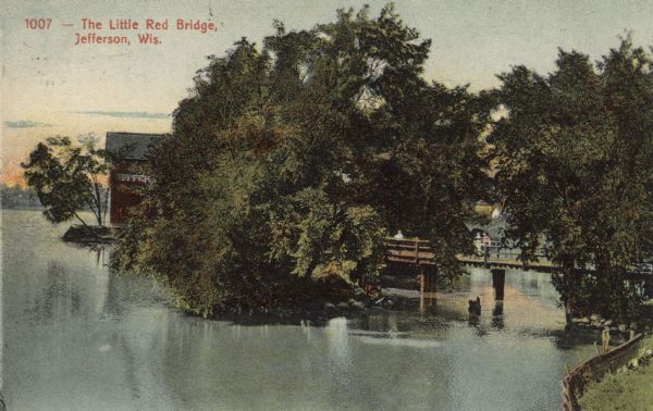 View from shoreline towards a footbridge across the Rock River. People are standing on the bridge. Caption reads: "The Little Red Bridge, Jefferson, Wis."
