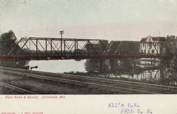 Hand-colored postcard view of a bridge over a river from railroad tracks. There is a building on the far shoreline. Caption reads: "Rock River and Bridge, Jefferson, Wis."