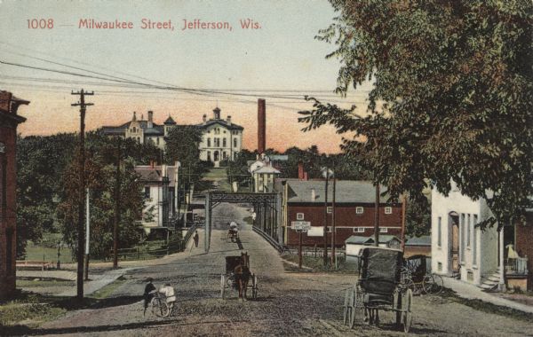 View of Milwaukee Street and the bridge crossing the Rock River. Horses and buggies are in the road. A building with a bell tower is in the background. Caption reads: "Milwaukee Street, Jefferson, Wis."