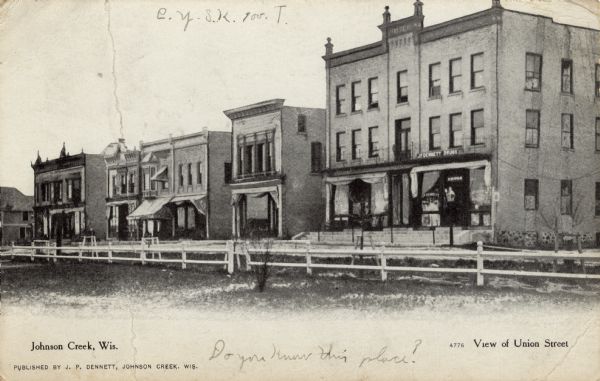 View across grass and fence towards the buildings along Union Street. J.P. Dennett Drugs is on the corner. Caption reads: "View of Union Street, Johnson Creek, Wis."