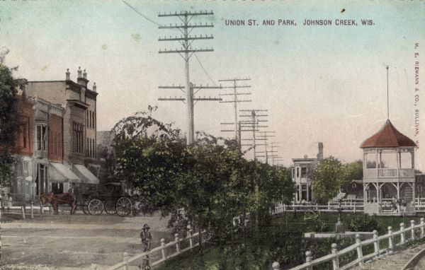 Elevated view of a park in the foreground with a fountain and a two-level gazebo on the right, and a row of buildings on the left. A horse and a wagon are parked in the street. Caption reads: "Union St. and Park, Johnson Creek, Wis."