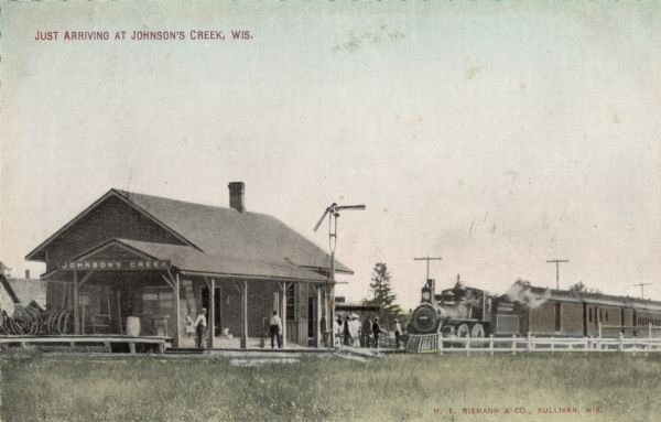 View of the Johnson Creek Depot and a train pulling up. Passengers are on the platform. Caption reads: "Just Arriving at Johnson's Creek, Wis."