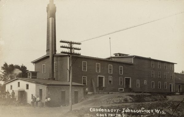 Photographic postcard view of the condensory. A group of workers are gathered outdoors on the left. Caption reads: "Condensory — Johnson Creek, Wis."