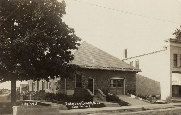 Photographic postcard view from street of the city hall. A grocery store is next door on the right. Caption reads: "City Hall, Johnson Creek, Wis."