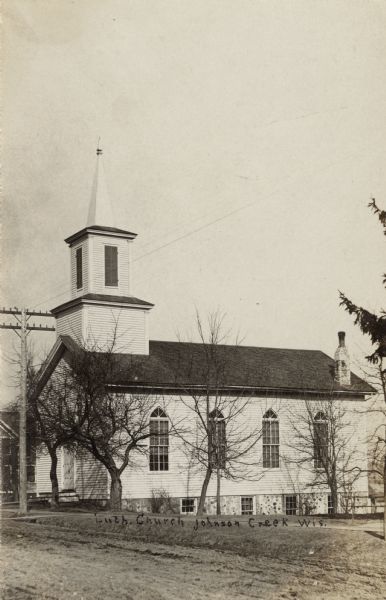 Photographic postcard view from unpaved road towards a wood-frame Lutheran church. There is a steeple and a bell tower above the entrance. Caption reads: "Luth. Church, Johnson Creek, Wis."
