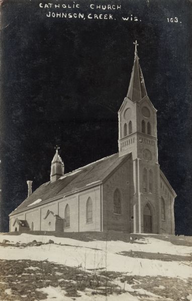 View of the Catholic church, with patchy snow on the lawn in front. There are two steeples, one in front and one in the rear. Caption reads: "Catholic Church, Johnson Creek, Wis."