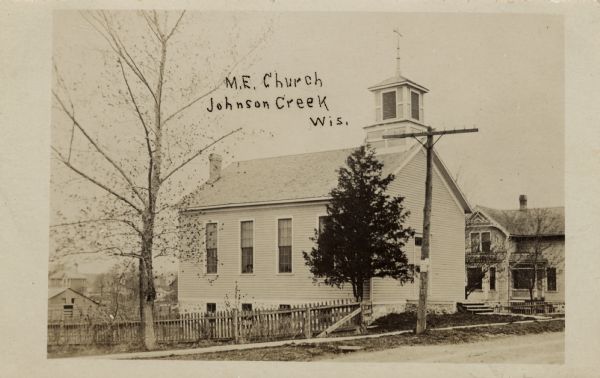 Photographic postcard view from unpaved road towards a small, wood-frame church with a fence on the left. There is a house next door on the right. Caption reads: "M.E. Church, Johnson Creek, Wis."