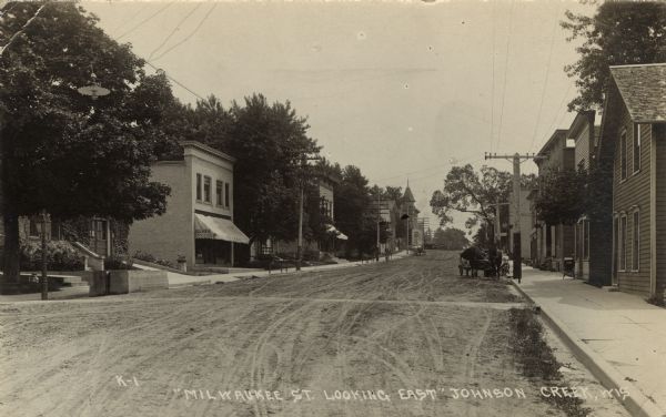 Photographic postcard view of a commercial district. There is a grocery store on the left, and horse-drawn wagons are along the curbs. Caption reads: "Milwaukee Street Looking East, Johnson Creek, Wis."