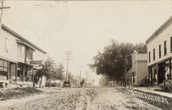 Photographic postcard view of a central business location. A drugstore is on the left, and a post office is on the right. A horse and buggy and an automobile are in the unpaved street. Caption reads: "Main Street Looking Southeast, Johnson Creek, Wis."