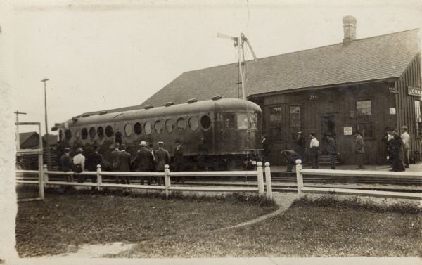 Photographic postcard view of a commuter rail car stopped at the depot. A group of businessmen are standing on the left near the railroad tracks, and more people are on the platform.