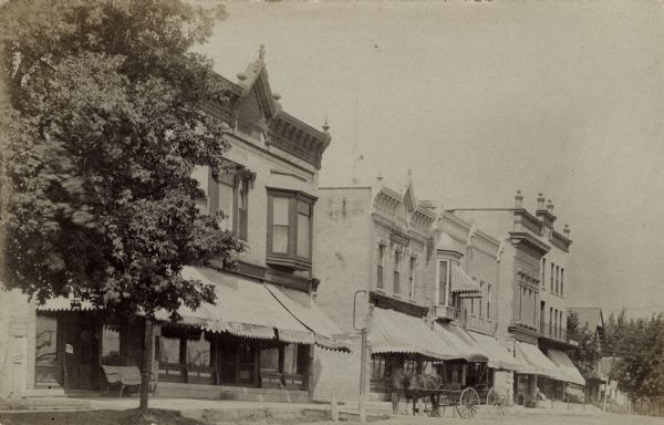 Photographic postcard view of a row of businesses in central Johnson Creek. A number of the storefronts have awnings above the show windows. A horse and buggy is parked at the curb.