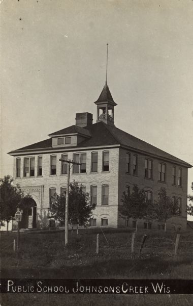 Photographic postcard view of the public school, a two-story brick building with a bell tower on the roof. Caption reads: "Public School, Johnson Creek, Wis."