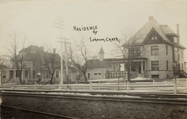 View of two homes with a stable in between. A fountain and railroad tracks are in the foreground. Caption reads: "Residence of Johnson Creek, Lake Mills, Wis."
