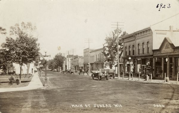 View of the businesses along Main Street. Automobiles and buggies are parked along the curb. The street is lined with street lights and hitching posts. Caption reads: "Main St., Juneau, Wis."