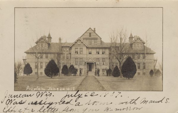 View towards a group of people standing on the grounds in front of the asylum. There are bars on the windows and fire escapes on both ends of the building. Caption reads: "Asylum, Juneau, Wis."