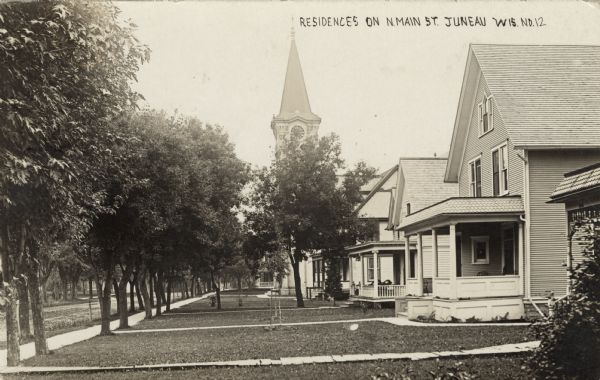 View of a row of homes with lawns and porches along Main Street. There is a church with a clock on the steeple in the background. Trees line the sidewalk along the left. Caption reads: "Residences on N. Main St. Juneau, Wis."