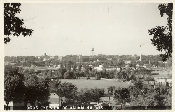 Photographic postcard of an elevated view of Kaukauna. There is a water tower in the center, a church on the left, and a smokestack on the right, and the Fox River passing through. Caption reads: "Birds [sic] Eye View of Kaukauna, Wis."